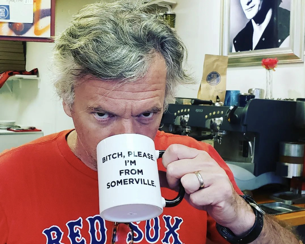 This photo shows Charlie Redd, with thick graying hair and wearing a Boston Red Sox shirt, sipping from a coffee cup emblazoned with the words, Bitch, please, I'm from Somerville