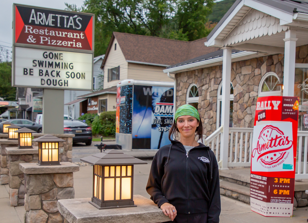 THis photo shows Erin McLaughlin standing in front of her pizzeria. Behind her is a sign that reads, Gone Swimming - Be Back Soon Dough