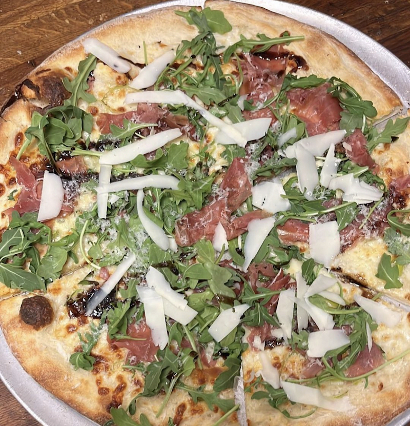This photo shows a white-sauce pizza topped with leeks, prosciutto, arugula and Asiago cheese.