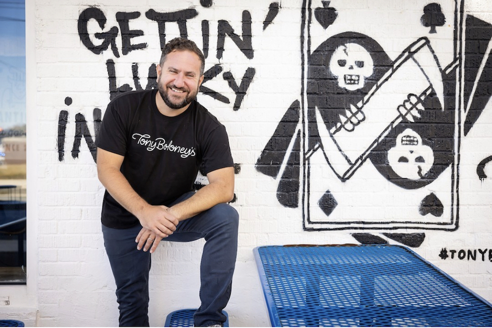 This photo shows Mike Hauke in a black Tony Boloney's t-shirt standing in front of a sign that seems to depict a twisted version of a playing card and the words "Getting Lucky In" something or another.