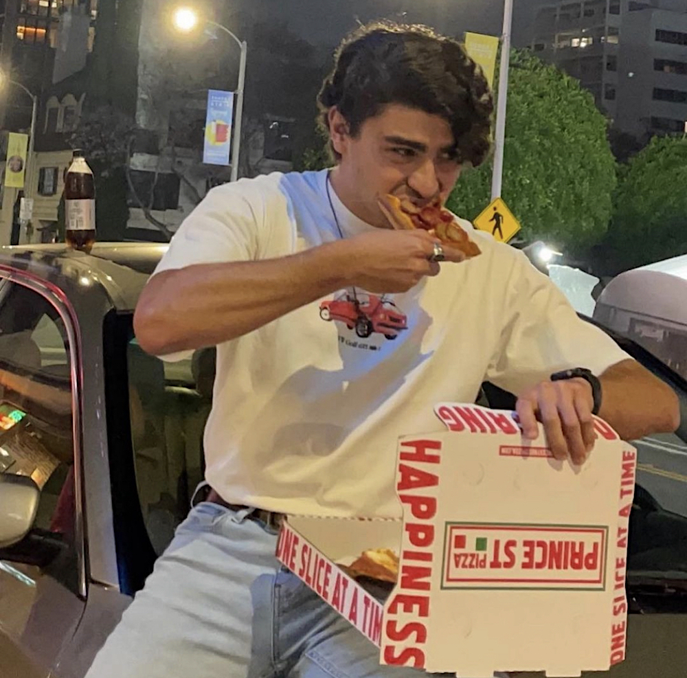 This photo shows a young man with thick, dark hair eating a slice of pizza outside from a Prince St. Pizza box.