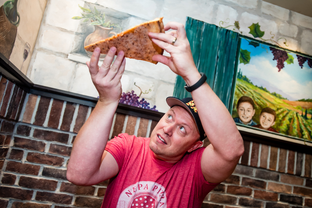 Here Jim is checking the bottom crust of a large slice of pizza. He's wearing a red NEPA Pizza Review t-shirt and a cap.