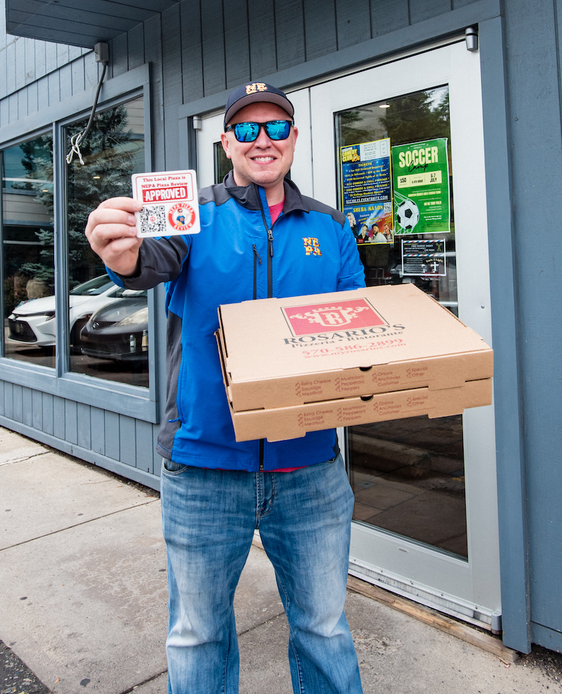 This photo shows Jim, wearing a blue jacket and blue sunglasses, holding up the NEPA Pizza Approved card in one hand and two carryout boxes of pizza from Rosario's