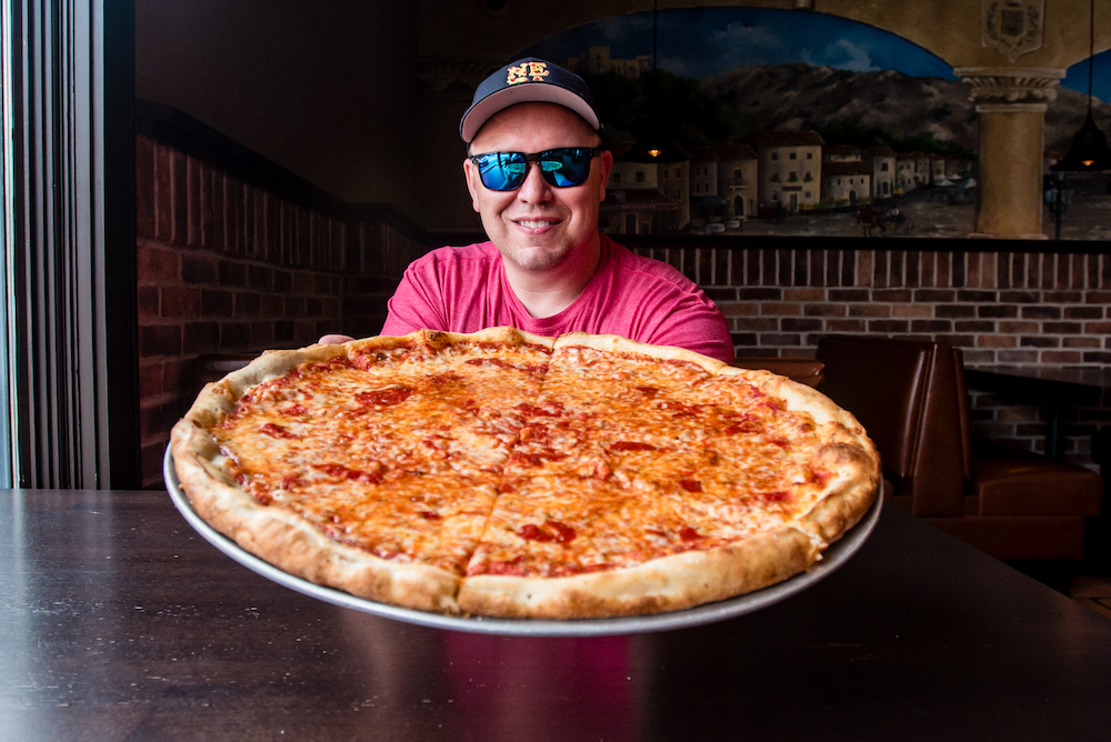 This photo shows Jim holding an extra large cheese pizza while wearing dark blue sunglasses, a cap and a red t-shirt.