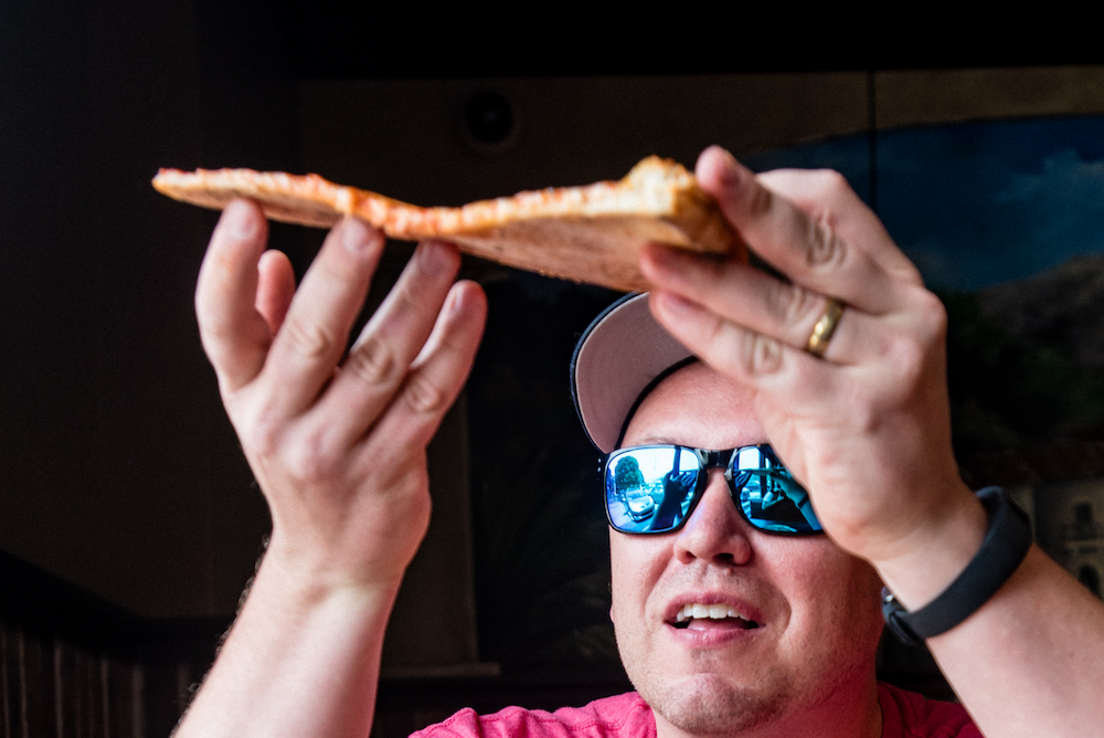 This photo shows Jim, wearing dark blue sunglasses, eyeing the undercarriage of a slice of pizza.