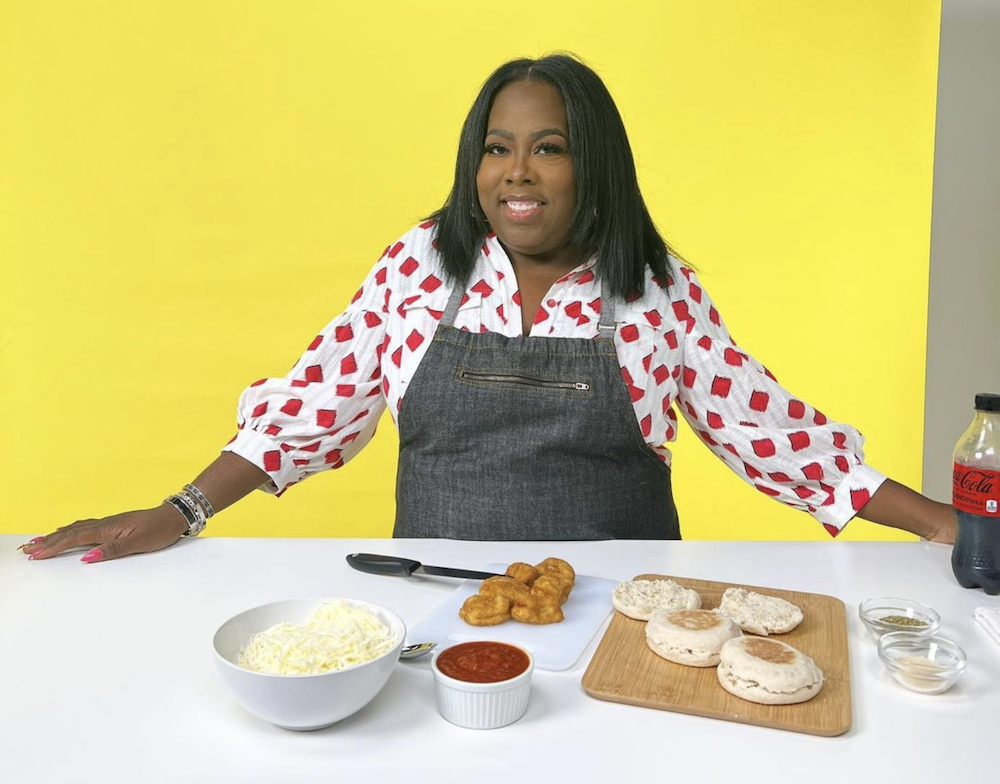 Photo shows a woman on the set of a cooking show with pizza ingredients in front of her.