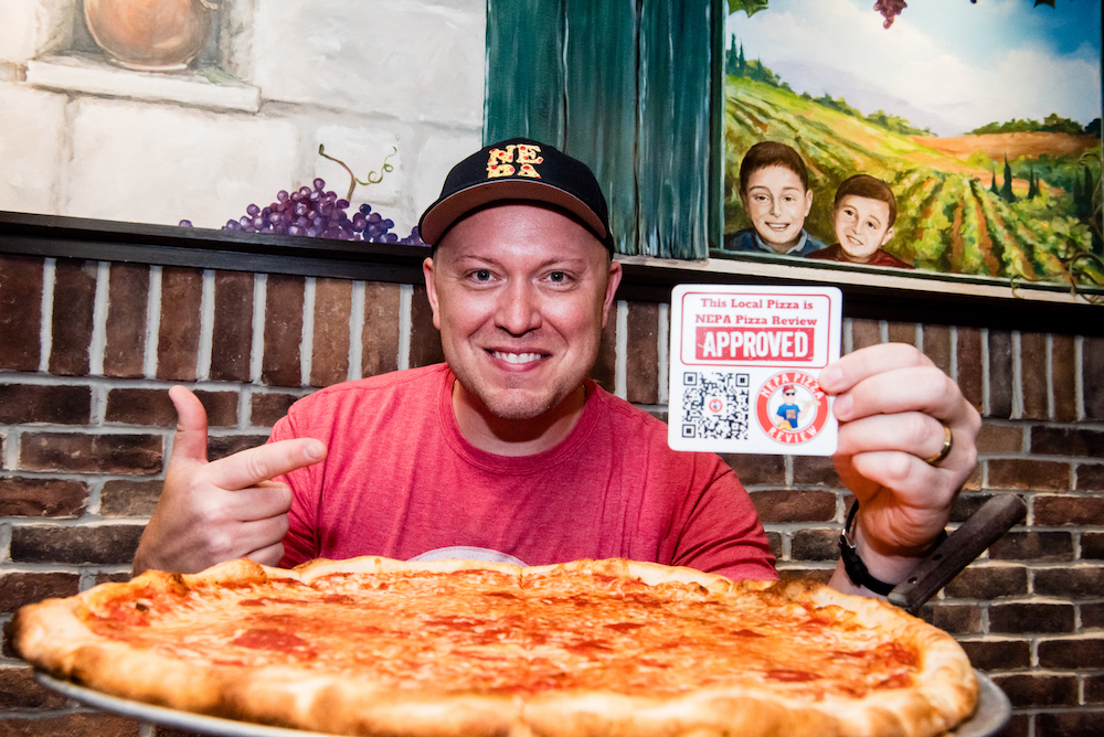 In this photo Jim Mirabelli is sitting behind a really large cheese pizza holding up a card that says This Local Pizza Is NEPA Pizza Approved