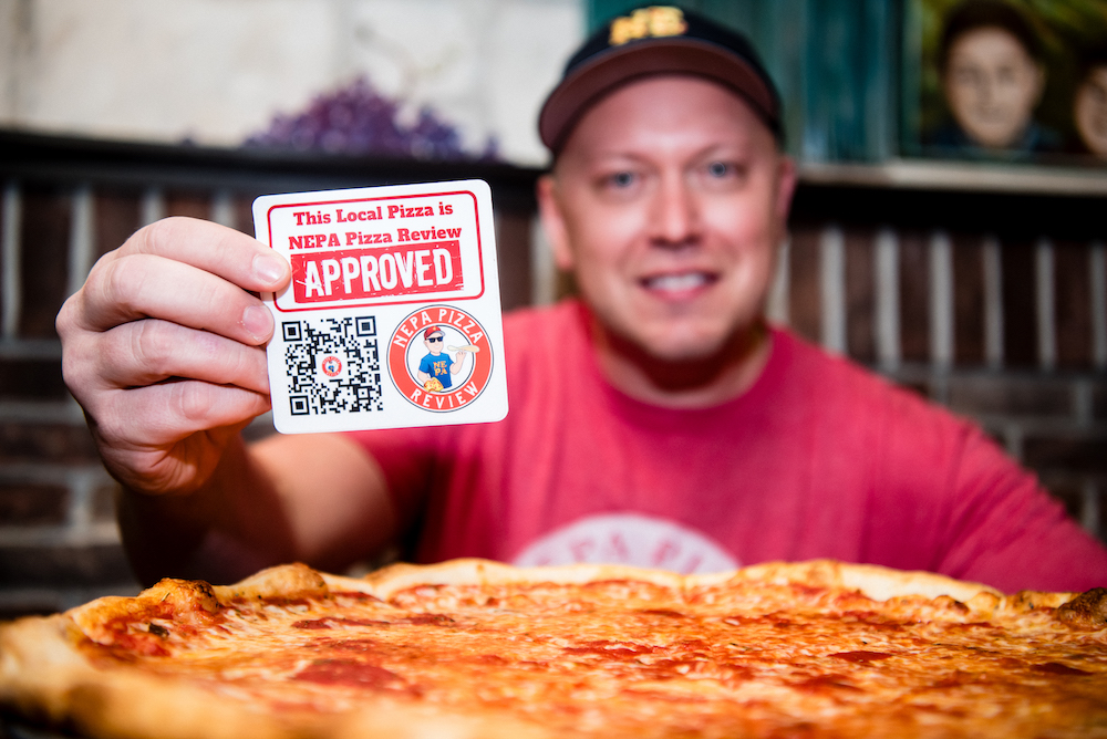 Here, Jim, wearing a red NEPA Pizza Review t-shirt, sits in front of a large cheese pizza and holds up a card that reads This Local Pizza is NEPA Pizza Approved.