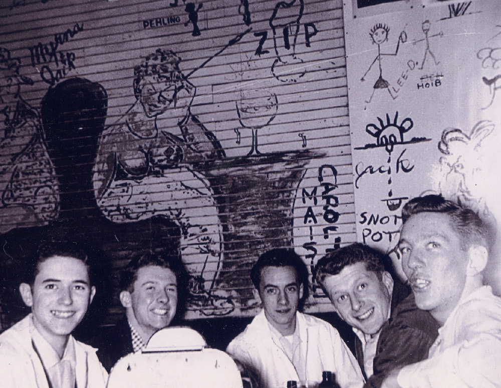 This vintage photo from the 1950s shows a group of happy-looking men seated in the restaurant in front of a wall decorated with various original artworks.