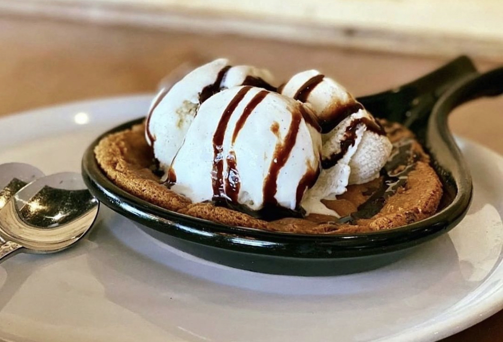 This photo shows a white plate with a chocolate chip cookie topped with vanilla ice cream and a chocolate drizzle.