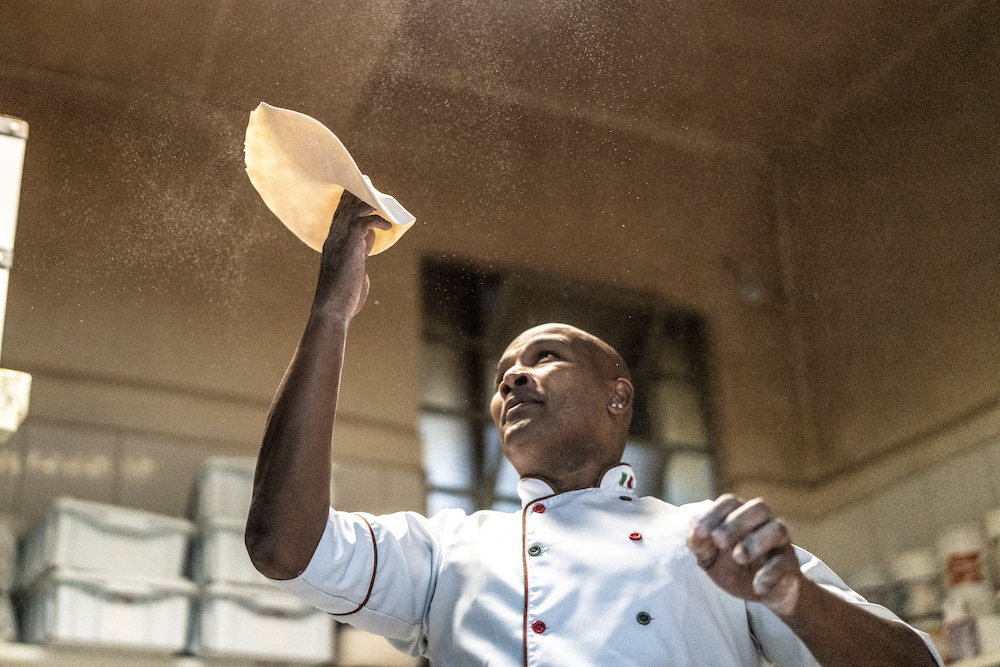 This photo shows a bald black pizza chef in a white chef coat spinning pizza dough.