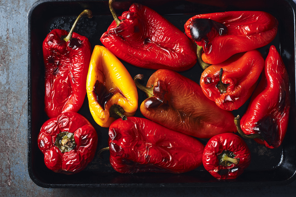 This photo shows a pan of roasted red peppers, nicely charred, with one yellow pepper on the left hand side.