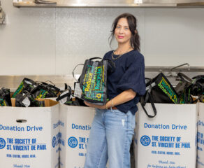 This photo shows Jaime Spinato, with long dark hair and wearing a dark blue shirt and blue jeans, holding a kindness bag and standing in front of boxes piled with the bags.