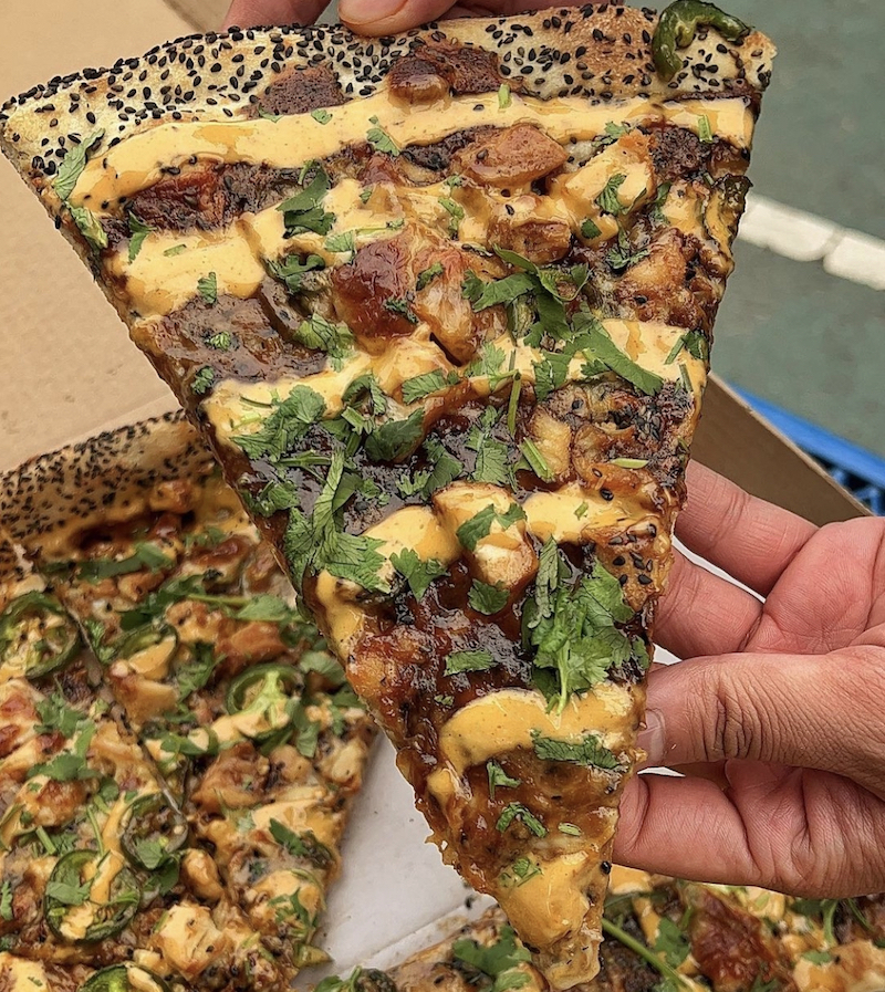 In this photo, the pizza slice has a sesame seed crust with Thai satay fried chicken, mozzarella, cilantro, peanuts and a yellowish sriracha Thai ranch dressing