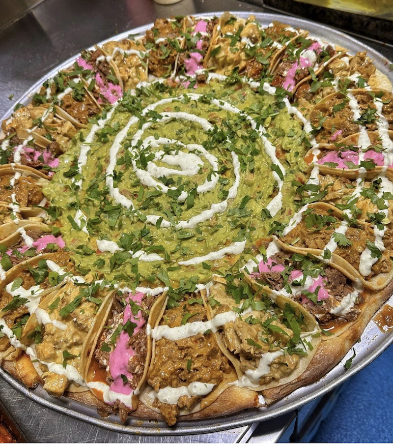 This photo shows a large pizza topped with a ring of mini-tacos and guacamole and sour cream at the center.