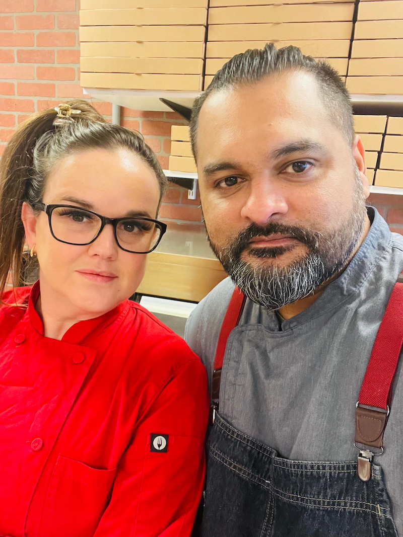 This photo shows Glaucia Franco, with her dark hair pulled back into a ponytail and wearing glasses and a red jacket, and Wesley Cavalavante, with a mustache and beard, wearing a gray shirt and dark-blue apron.