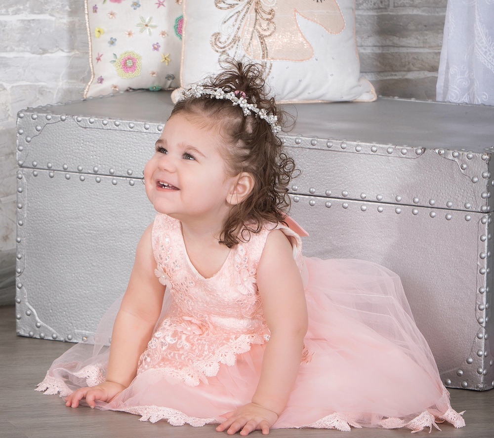 This photo shows the adorable three-year-old Collyns on her knees, wearing a pink princess dress and a tiara, looking away from the camera and smiling sweetly.