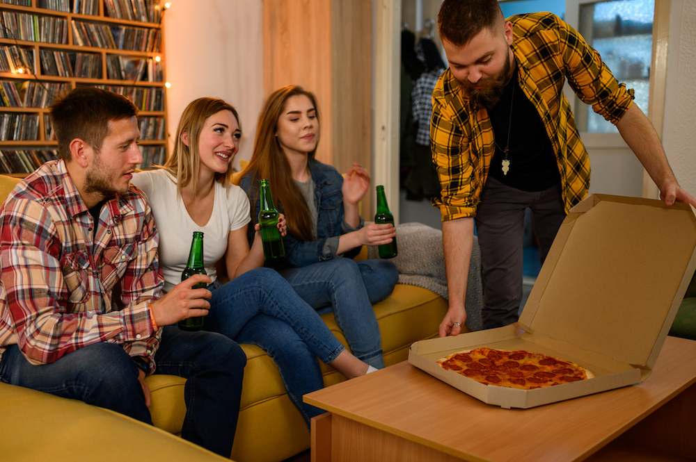 Group of friends eating pizza on a house party and having fun while drinking beer an sitting on the couch all together