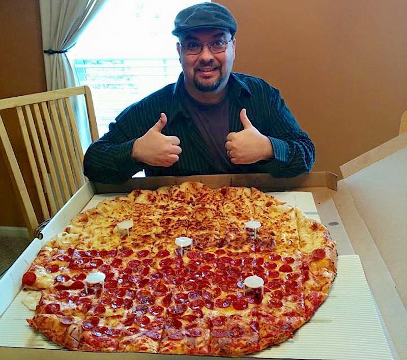 This shows a man wearing a dark cap and striped dark shirt giving the thumbs-up as he sits in front of the gigantic Tarantosaurus pizza.