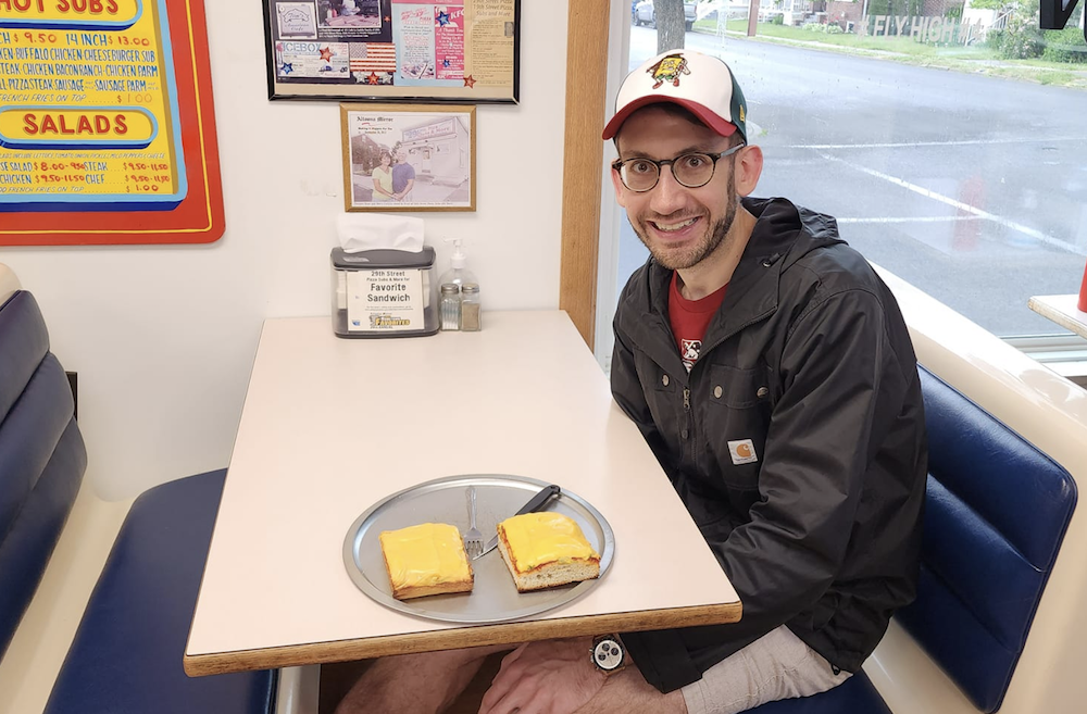 A man sits donning an Altoona-style pizza-themed minor league baseball hat, about to dig into his altoona-style pizza.