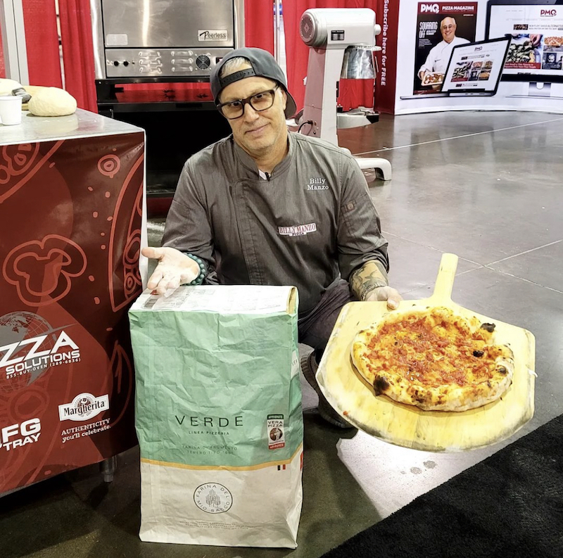 This photo shows a man wearing a backwards baseball cap and wearing a gray chef's coat, holding a pizza peel with a huge baked pizza on it and standing in front of what looks to be a huge bag of flour