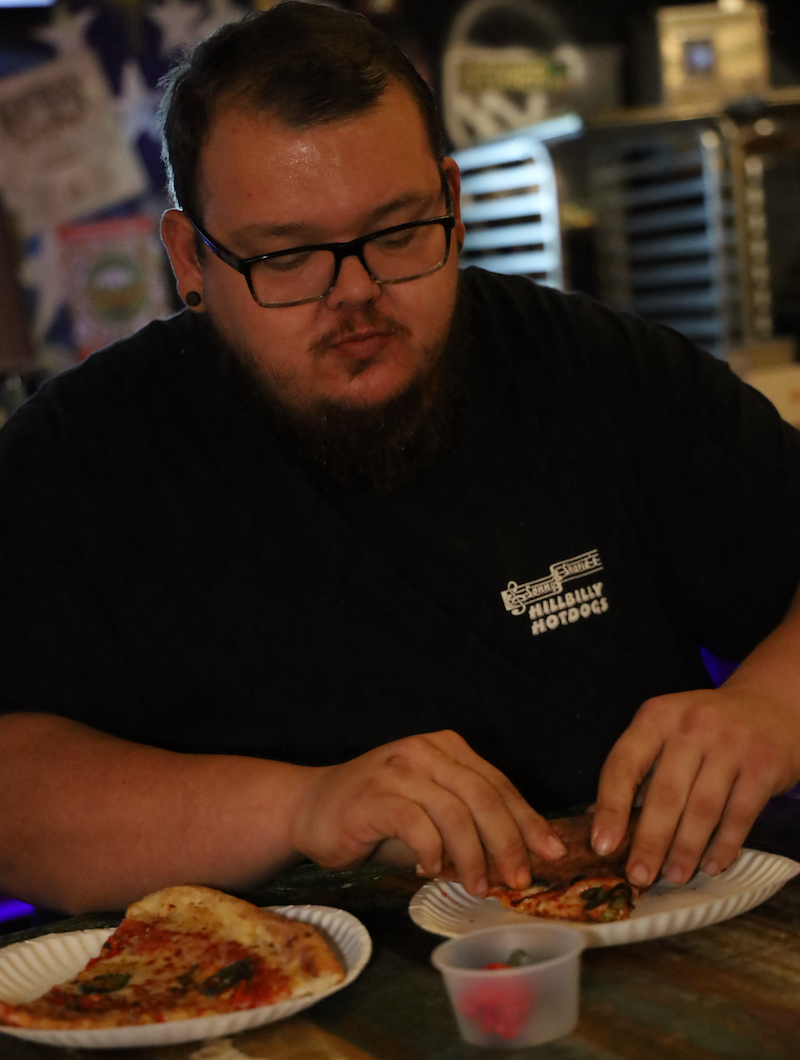 This photo shows the contest winner, with short dark hair, glasses and a black t-shirt, eating pizza.