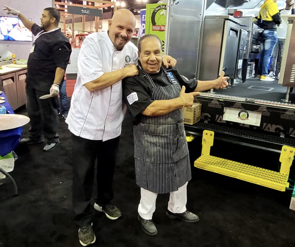 This photo shows two men, one in a white chef coat and a much shorter man in a gray chef coat, pointing to a truck that's loaded with pizzeria equipment.