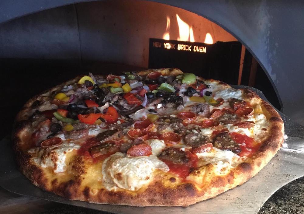 This photo shows a pizza topped with various meats and veggies positioned in front of a wood-fired oven with the flames burning in the background.