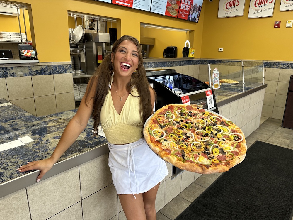 This photo shows Giuliana Calascibetta holding a huge pizza, wearing a yellow sleeveless shirt and a white skirt