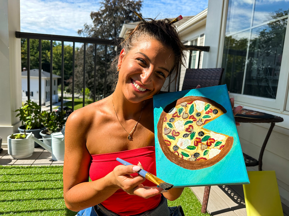 This photo shows a smiling Giuliana outdoors on a sunny day, holding up a painting of a pizza with one slice cut out.