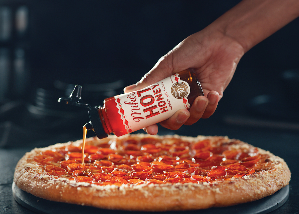 This photo shows a woman's hand holding a bottle of Mike's Hot Honey and drizzling it over the Pepperoni Magnifico pizza.