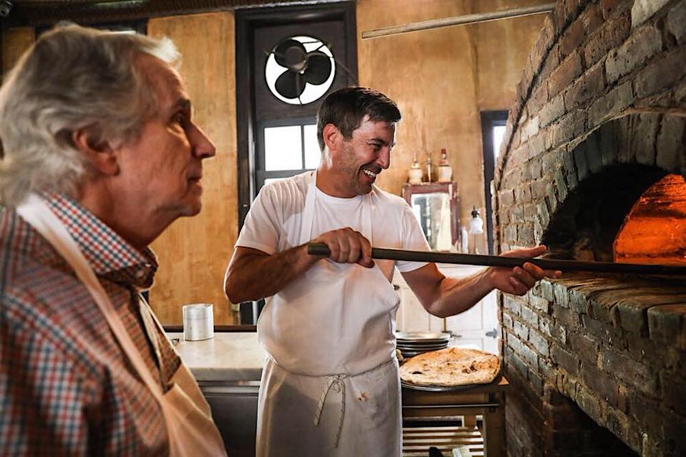 This shows Mark Iacono peeling a pizza into his oven and smiling while an unidentified older gentleman with gray hair looks on.