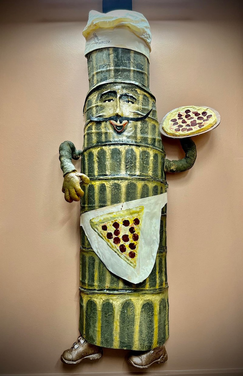 This photo shows a green and yellow, cylindrical sculpture of an Old World Italian pizza man with mustache and a chef's hat.