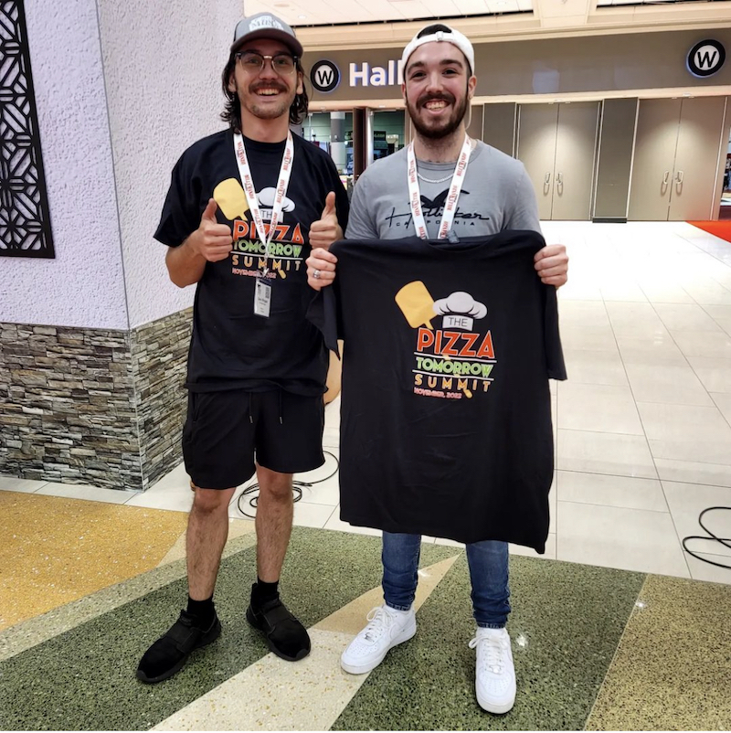 This photo shows two men, one wearing a backwards baseball cap and the other wearing his cap forward, posing with Pizza Tomorrow Summit t-shirts