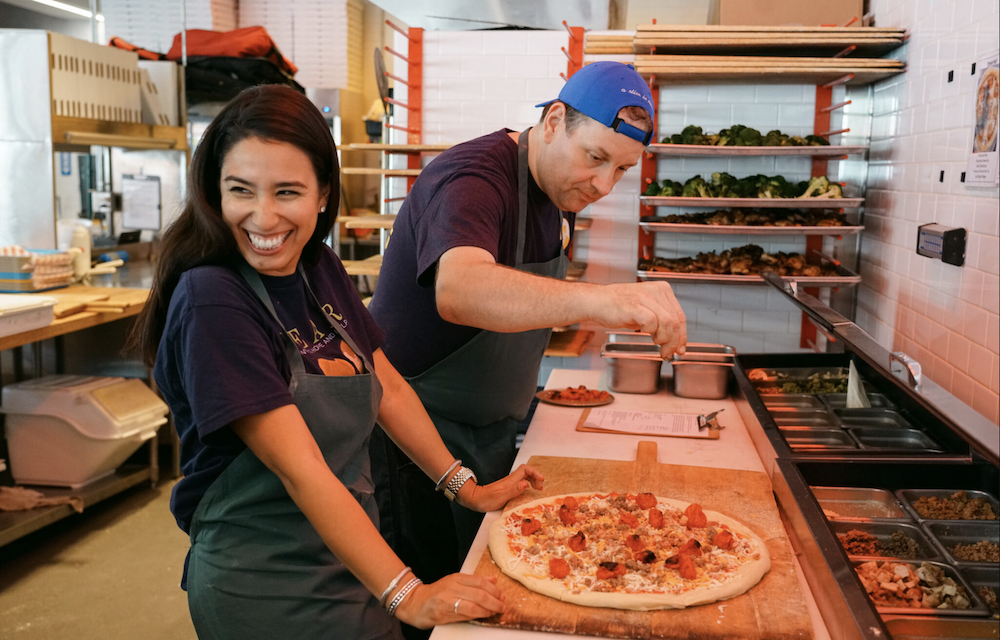 Vinny's Pizza executive chef Paul Lewis sprinkles ingredients atop a pie as a member of BEAR, an organization in Houston that helps neglected children, smiles alongside him.