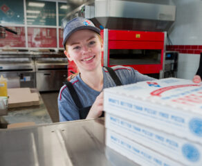 This photo shows a young woman wearing a blue Domino's cap and shirt placing several carryout pizza boxes on a counter and smiling.