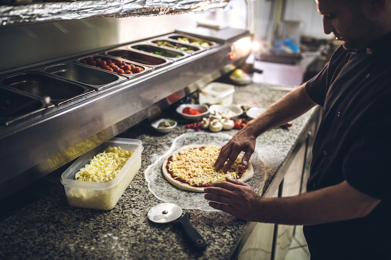 This photo shows a male chef adding cheese to an unbaked pizza crust.