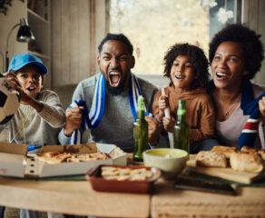 This photo shows a cheerful African American family watching a successful soccer game on TV while eating pizza at home.