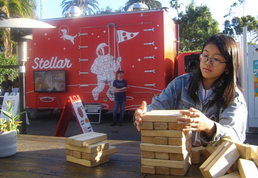 This photo shows a woman stacking what looks like pizzas of wood, with the Stellar Pizza truck parked behind her.