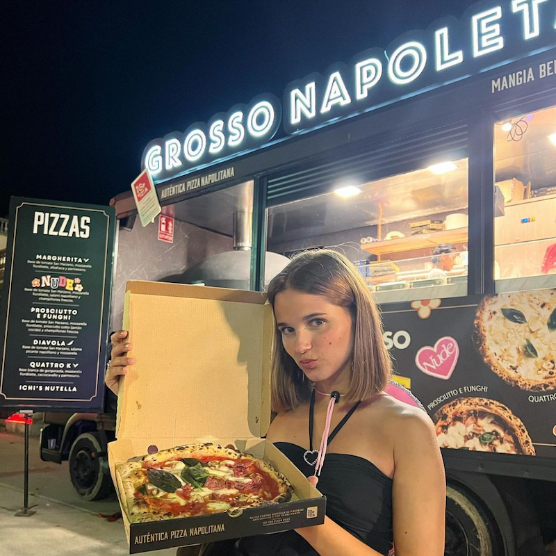 This photo shows an attractive young woman standing outside Grosso Napoletana, pursing her lips and holding up an open box with a pizza inside it.