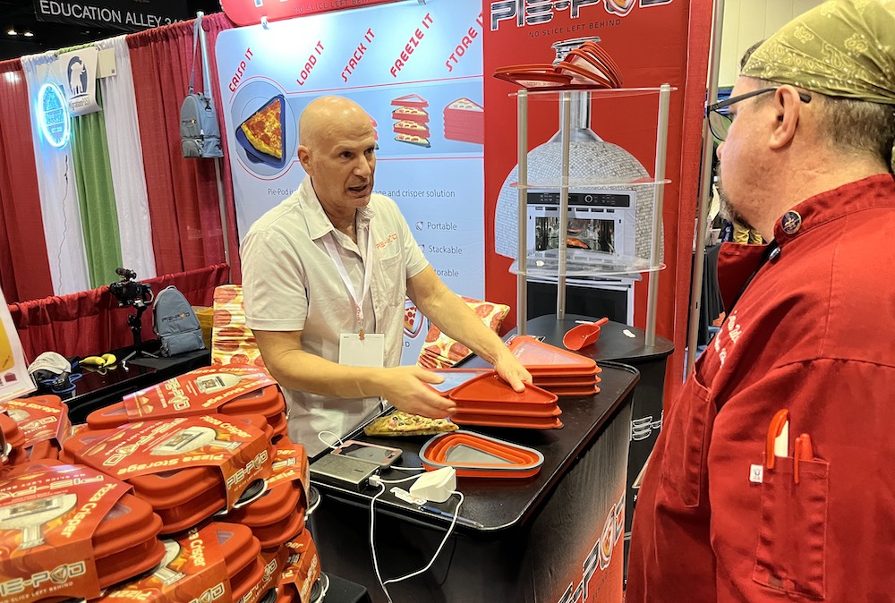 This photo shows a rep from Pie Pod demonstrating how their product, a red storage container for individual pizza slices, works.