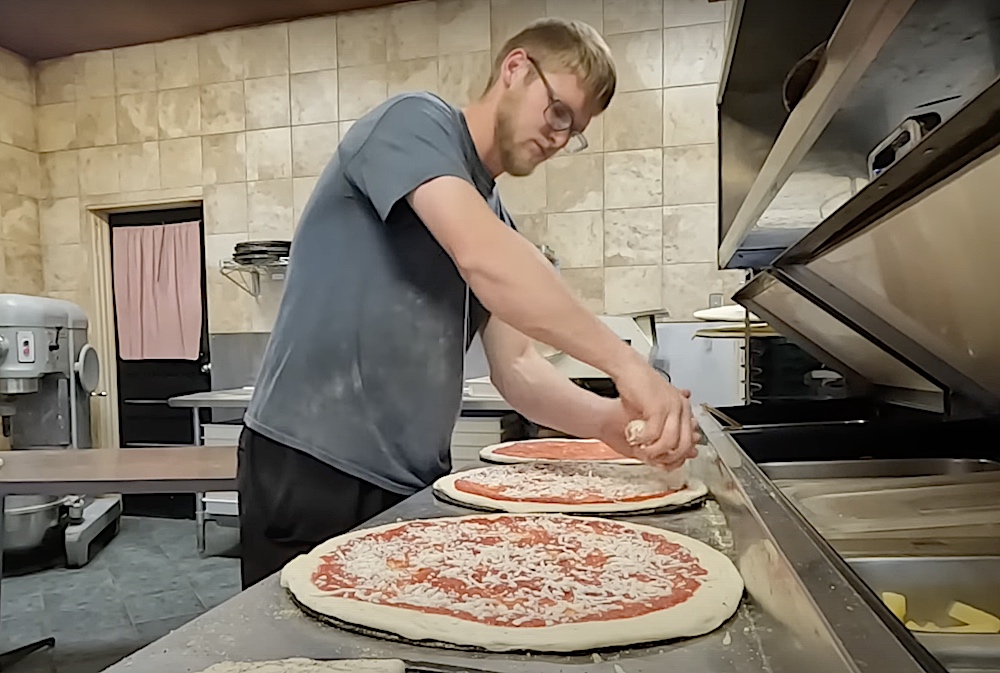 This photo shows Collin Brackin, with blondish hair and a beard and wearing a gray t-shirt, adding cheese to a pizza crust before baking it.