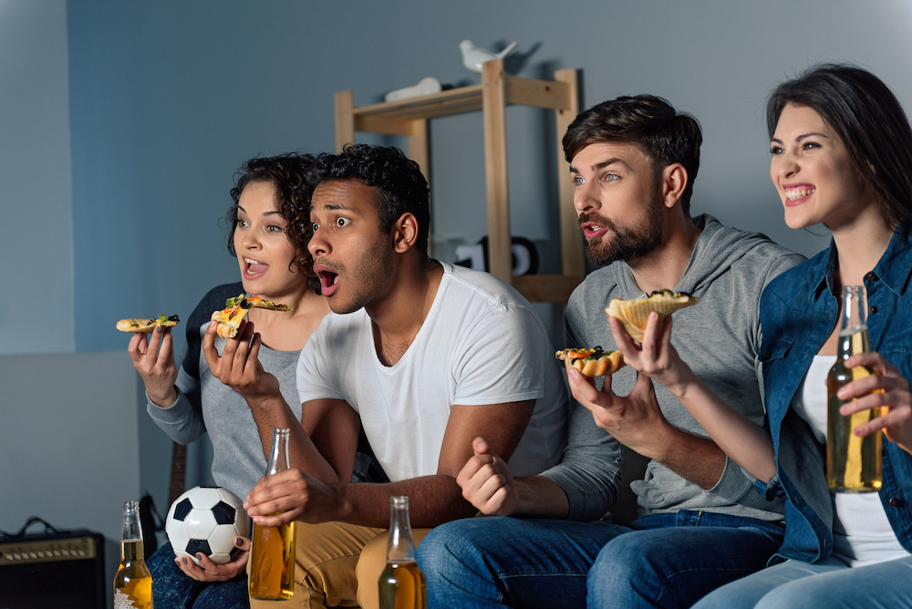 This photo shows a diverse group of sports fans eating pizza and cheering for their team as they watch a game on TV.