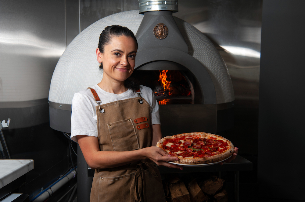 This photo shows Marisol Doyle standing in front of a wood-burning oven, smiling and displaying a beautiful pepperoni pizza