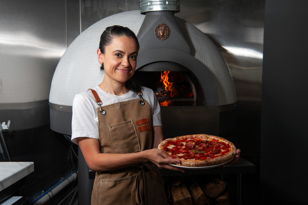 This photo shows Marisol Doyle standing in front of a wood-burning oven, smiling and displaying a beautiful pepperoni pizza