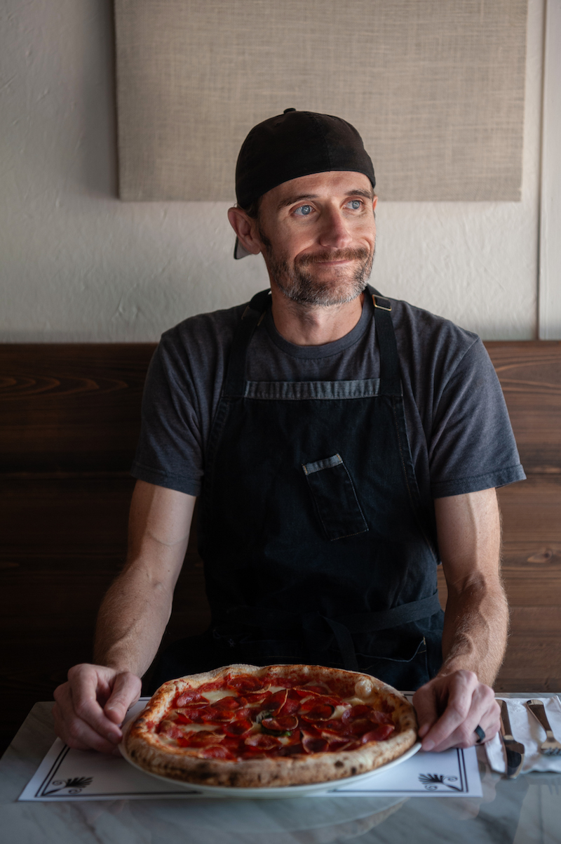 This photo shows Rory Doyle, wearing a backwards cap, a dark gray t-shirt and a dark apron, sitting in Leña with a pepperoni pizza in front of him.