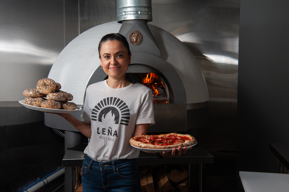 This photo shows Marisol Doyle in a white Lena t-shirt, standing in front of a wood-burning oven. She's holding a tray of a beautiful Neapolitan pepperoni pizza in one hand and a tray of bagels in the other.