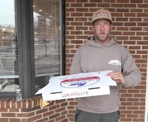 This photo shows Dave Portnoy, wearing a grey sweatshirt and cap, holding a pizza box from Lucatelli's and standing in front of the restaurant.