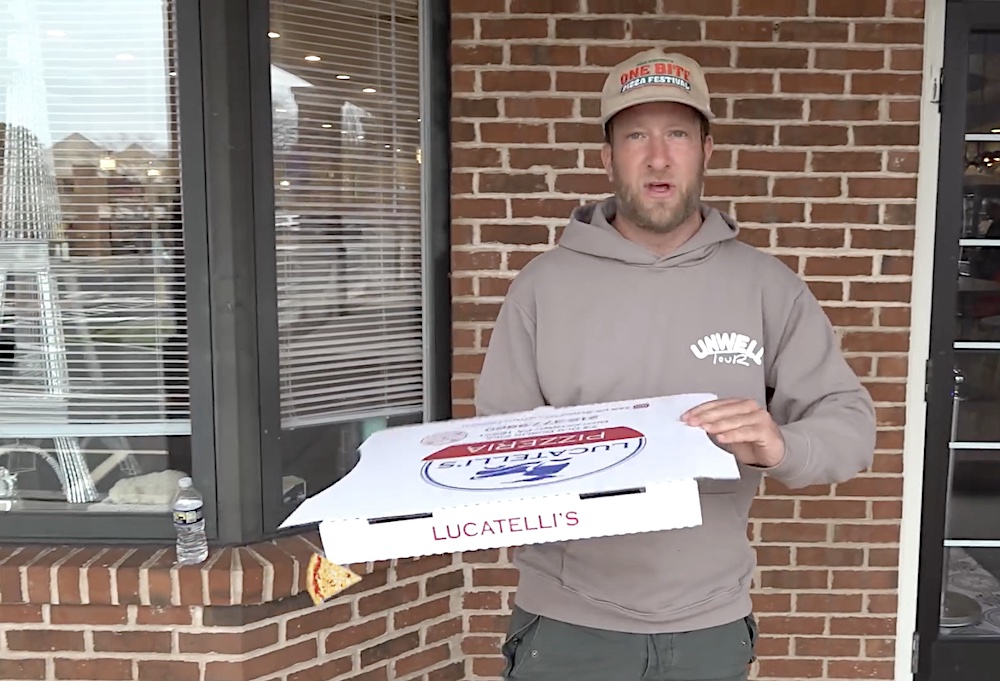 This photo shows Dave Portnoy, wearing a grey sweatshirt and cap, holding a pizza box from Lucatelli's and standing in front of the restaurant.