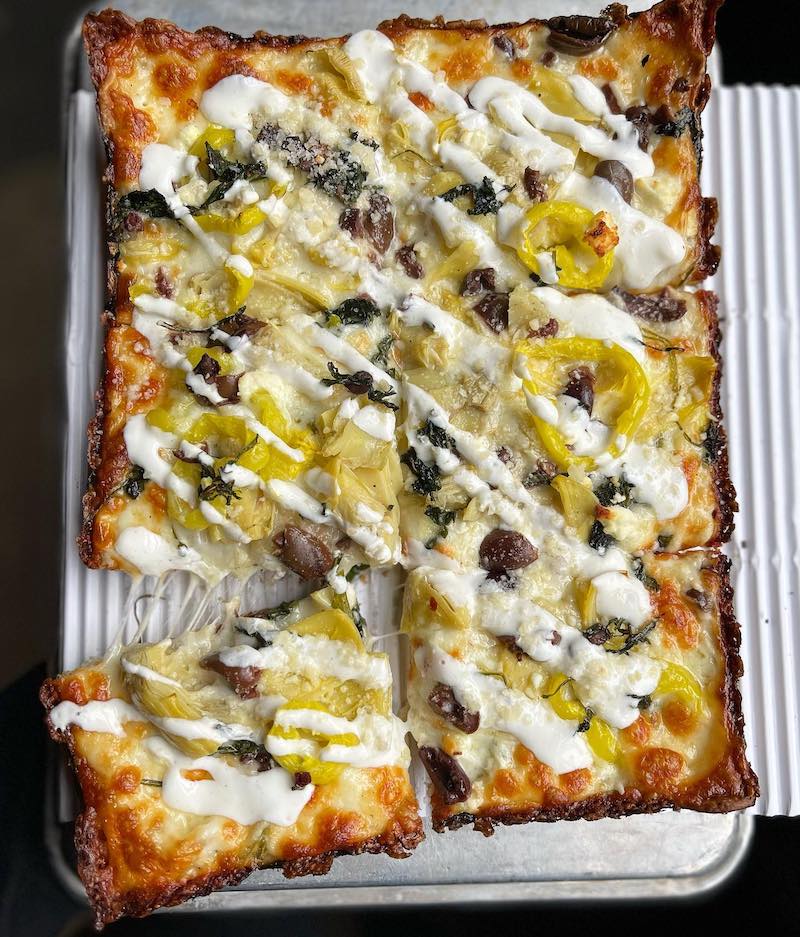 This photo shows a Detroit-style pizza topped with mozzarella, feta, artichokes, banana peppers, Kalamata olives and roasted garlic labneh.